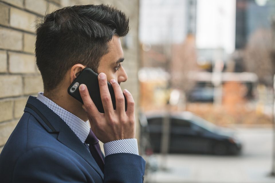 A young man in a navy suit and checked shirt taking a phone call in front of a brick wall